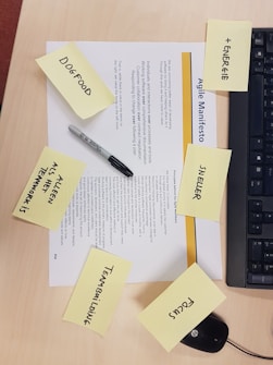 A printed document titled 'Agile Manifesto' is placed on a wooden desk. Surrounding the document are several yellow sticky notes inscribed with words like 'Dogfood,' '+ ENERGIE,' 'NEUER,' 'Focus,' 'TEAMBUILDING,' and 'ALS KLEEN TEAMWORK IS.' A black Sharpie pen and part of a black computer keyboard are also visible.