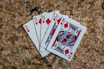 A hand of five playing cards is spread out on a textured, stone-like surface. The cards are of the diamond suit, including the 2, 5, 6, Jack, and King. The face cards feature intricate designs with red, black, and blue colors.