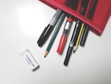 A collection of writing and drawing tools is partially placed inside a red pencil case. Items include a Sharpie marker, a red pen, pencils, a mechanical pencil, and an eraser lying nearby on a plain surface.