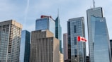 A skyline featuring modern skyscrapers with varied architectural designs, including logos of major corporations such as KPMG, BMO, and EY. A prominent Canadian flag is waving in the foreground. The sky is mostly clear, contributing to a well-lit scene.