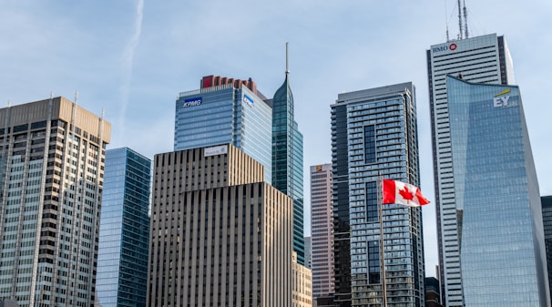 A skyline featuring modern skyscrapers with varied architectural designs, including logos of major corporations such as KPMG, BMO, and EY. A prominent Canadian flag is waving in the foreground. The sky is mostly clear, contributing to a well-lit scene.