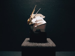 A model of a robot or mechanical figure is prominently displayed on a textured black pedestal against a dark background. The figure is predominantly white with some yellow and grey accents. The scene is dramatically lit, casting shadows and highlighting the contours of the object.