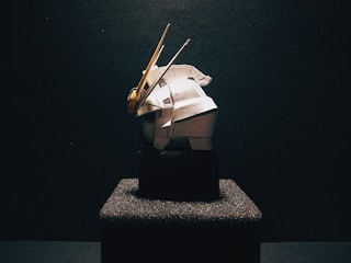 A model of a robot or mechanical figure is prominently displayed on a textured black pedestal against a dark background. The figure is predominantly white with some yellow and grey accents. The scene is dramatically lit, casting shadows and highlighting the contours of the object.