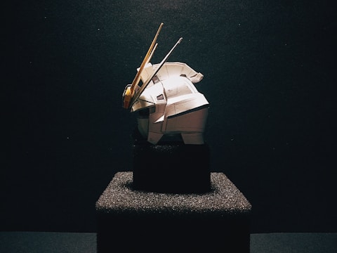 A model of a robot or mechanical figure is prominently displayed on a textured black pedestal against a dark background. The figure is predominantly white with some yellow and grey accents. The scene is dramatically lit, casting shadows and highlighting the contours of the object.