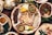 A variety of traditional dishes are arranged on a wooden table. The central platter contains slices of sausage, fried pork rinds, grilled meat, and various dipping sauces. Surrounding it are additional dishes with spices, vegetables, and garnishes, creating a colorful and appetizing display.