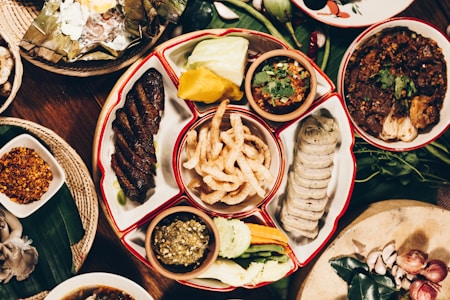 A variety of traditional dishes are arranged on a wooden table. The central platter contains slices of sausage, fried pork rinds, grilled meat, and various dipping sauces. Surrounding it are additional dishes with spices, vegetables, and garnishes, creating a colorful and appetizing display.
