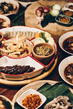 A variety of traditional Thai dishes is displayed on a wooden table. The centerpiece features a divided platter with deep-fried pork rinds, sliced sausages, mixed fresh vegetables, and a green chili dip. Surrounding this are various small bowls containing different sauces, spices, and broths. Raw ingredients, like shallots and green chilies, are visible on a wooden cutting board in the background.