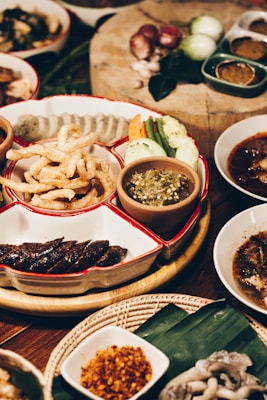 A variety of traditional Thai dishes is displayed on a wooden table. The centerpiece features a divided platter with deep-fried pork rinds, sliced sausages, mixed fresh vegetables, and a green chili dip. Surrounding this are various small bowls containing different sauces, spices, and broths. Raw ingredients, like shallots and green chilies, are visible on a wooden cutting board in the background.