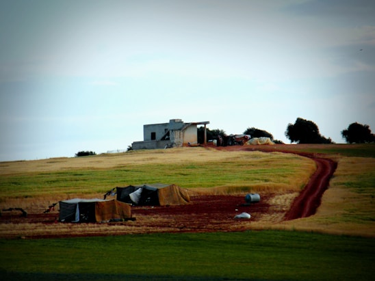 A rural landscape featuring a small structure atop a gentle hill surrounded by earthy, agricultural fields. The foreground includes a few tents or tarpaulin-covered structures and a red dirt path winding towards the building. Scattered trees dot the horizon, under a partly cloudy sky.