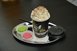 A hot beverage in a black paper cup topped with whipped cream and cocoa powder. The cup is placed on a shiny, round metal tray alongside two colorful macarons, one green and one pink, in white paper cups. A black plastic lid and a small spoon are also on the tray, resting on a white napkin with red text.