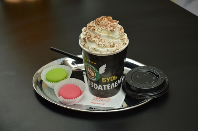 A hot beverage in a black paper cup topped with whipped cream and cocoa powder. The cup is placed on a shiny, round metal tray alongside two colorful macarons, one green and one pink, in white paper cups. A black plastic lid and a small spoon are also on the tray, resting on a white napkin with red text.