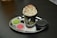 A hot beverage in a black paper cup topped with whipped cream and cocoa powder. The cup is placed on a shiny, round metal tray alongside two colorful macarons, one green and one pink, in white paper cups. A black plastic lid and a small spoon are also on the tray, resting on a white napkin with red text.