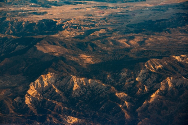 An aerial view of a rugged mountainous landscape with varying elevations and ridges. The sunlight casts a warm glow over the terrain, highlighting the textures and contours of the land.
