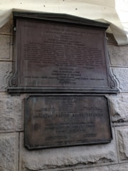 Two bronze plaques affixed to a rough stone wall. The upper plaque is larger, containing multiple lines of text including names and titles. The lower plaque is also text-heavy, mentioning the Brooklyn Bridge Reconstruction, the Department of Public Works, and various individuals involved.