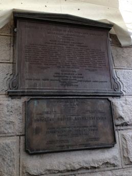 Two bronze plaques affixed to a rough stone wall. The upper plaque is larger, containing multiple lines of text including names and titles. The lower plaque is also text-heavy, mentioning the Brooklyn Bridge Reconstruction, the Department of Public Works, and various individuals involved.