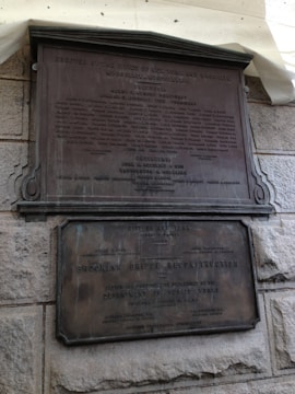 Two bronze plaques affixed to a rough stone wall. The upper plaque is larger, containing multiple lines of text including names and titles. The lower plaque is also text-heavy, mentioning the Brooklyn Bridge Reconstruction, the Department of Public Works, and various individuals involved.