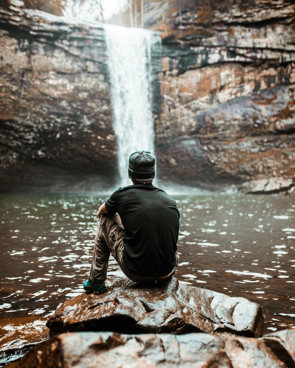 man sitting on brown rock near waterfalls at day time