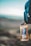 A reusable plastic water bottle with a bear sticker is attached to a dark-colored backpack. The background is blurred, suggesting an outdoor environment possibly with a sky and landscape.