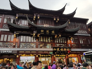 A traditional Chinese architectural building with ornate, curved roofs and intricate wooden detailing serves as a backdrop. The building has signs with Chinese characters and the words 'Song Yun Tower.' Below, a bustling crowd of people is visible, indicating a busy street scene with various shops, including a coffee shop.