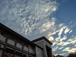 A building with multiple windows and a sign that reads 'BANK INDONESIA' is depicted under a sky filled with scattered clouds. The clouds are illuminated by sunlight, creating a dynamic contrast with the blue sky.
