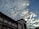 A building with multiple windows and a sign that reads 'BANK INDONESIA' is depicted under a sky filled with scattered clouds. The clouds are illuminated by sunlight, creating a dynamic contrast with the blue sky.