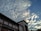 A building with multiple windows and a sign that reads 'BANK INDONESIA' is depicted under a sky filled with scattered clouds. The clouds are illuminated by sunlight, creating a dynamic contrast with the blue sky.