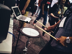 A person is holding drumsticks near an electronic drum pad connected to a laptop. The setup seems to be part of a music demonstration or trade show. Other people are visible in the background, suggesting a public event or exhibition.