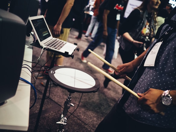 A person is holding drumsticks near an electronic drum pad connected to a laptop. The setup seems to be part of a music demonstration or trade show. Other people are visible in the background, suggesting a public event or exhibition.