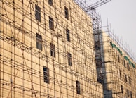 A building under construction is covered with an extensive network of metal scaffolding. The structure is beige with evenly spaced windows, and a crane is visible in the background.