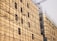 A building under construction is covered with an extensive network of metal scaffolding. The structure is beige with evenly spaced windows, and a crane is visible in the background.