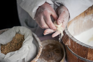 Hands working with thin strands of dough accompanied by a bowl with a powdery substance, possibly flour, and a large bag containing seeds. The environment suggests a focus on intricate food preparation.