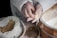 Hands working with thin strands of dough accompanied by a bowl with a powdery substance, possibly flour, and a large bag containing seeds. The environment suggests a focus on intricate food preparation.