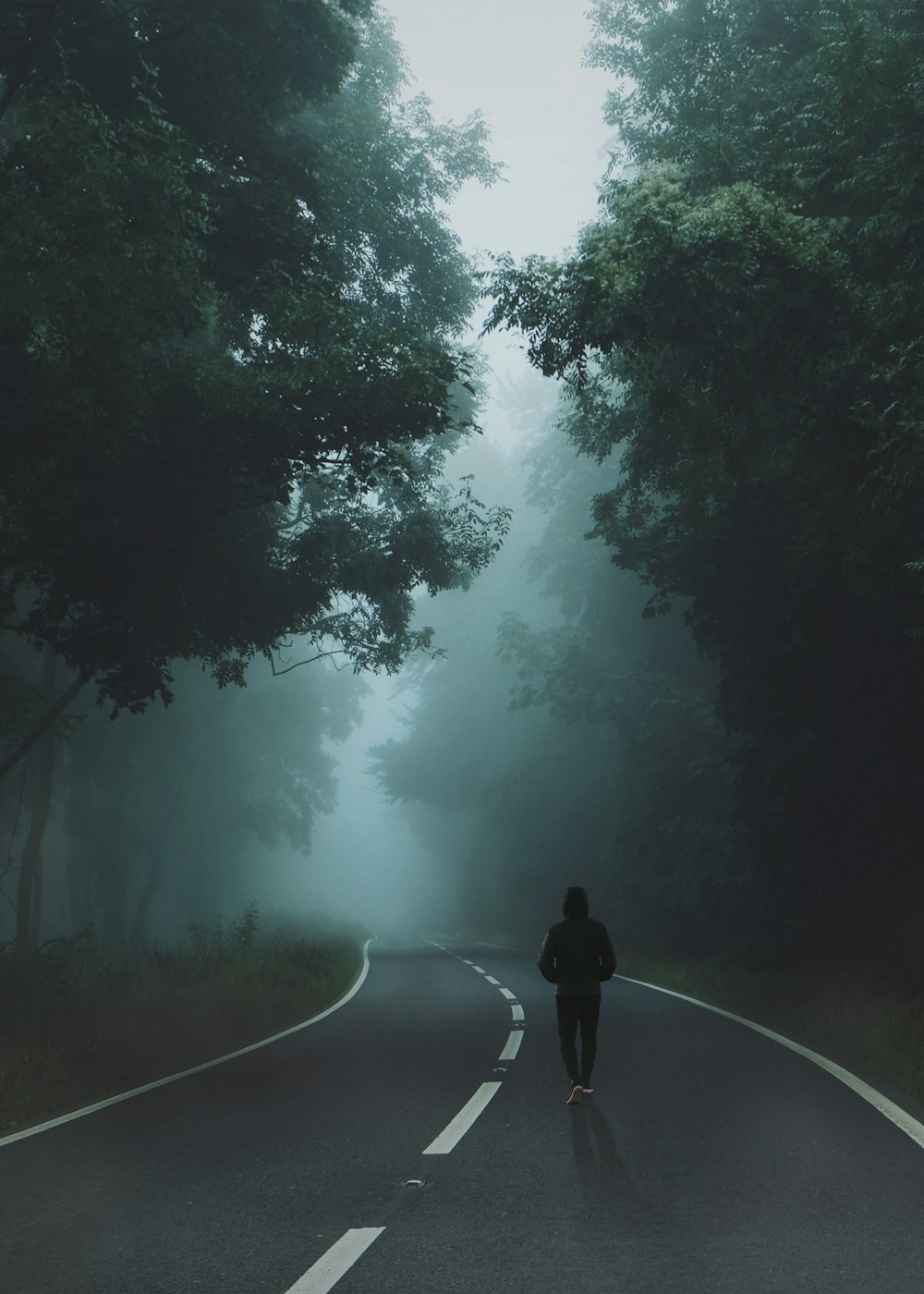 Hombre con sudadera con capucha negra y pantalones caminando por la carretera superior gris