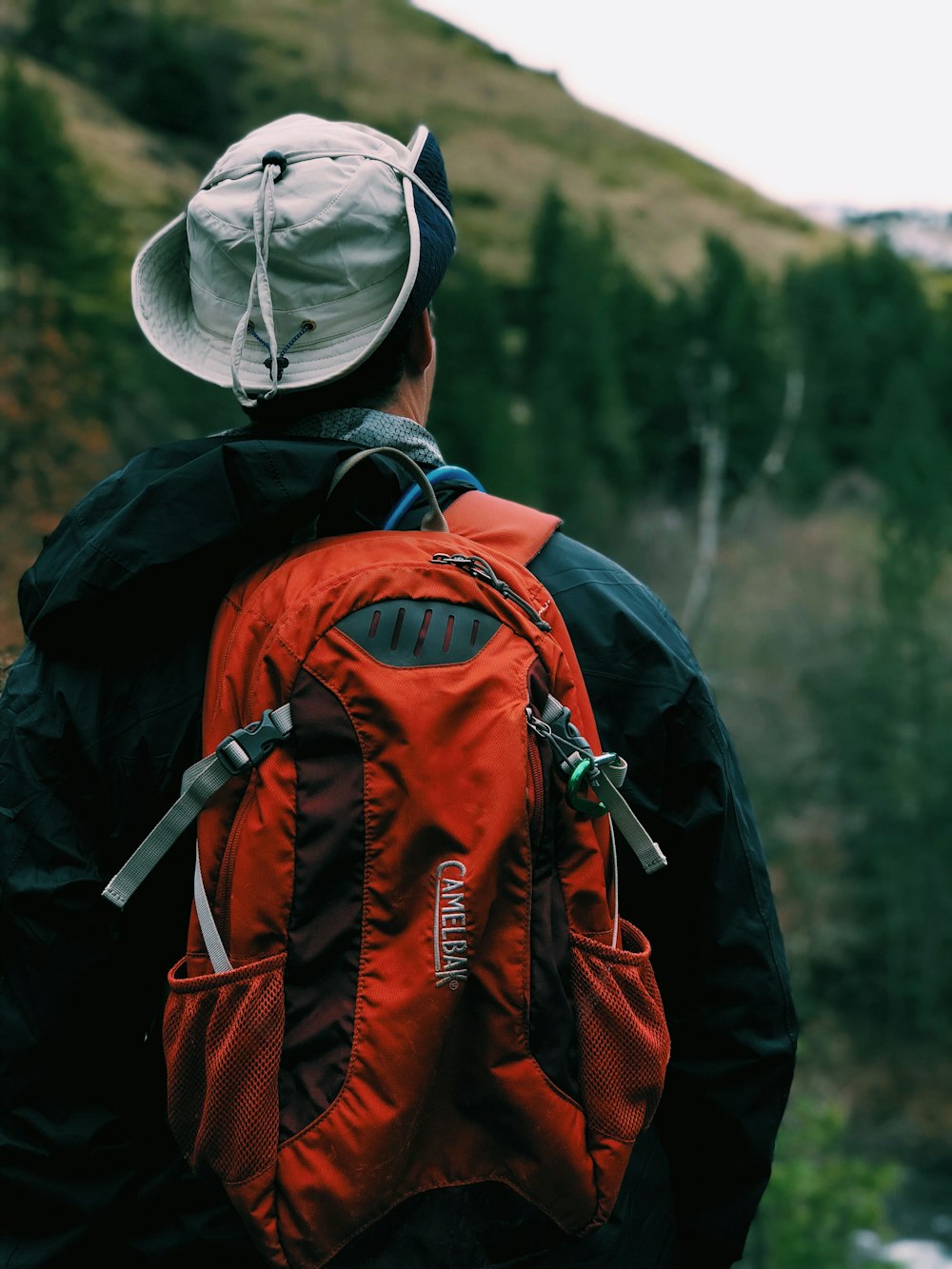 homem de casaco preto usando mochila vermelha em pé na frente da montanha durante o dia