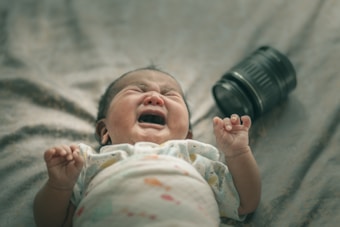 A crying baby lies on a soft surface, dressed in a light-colored outfit with a pattern. Next to the baby is a black camera lens lying on its side.