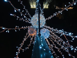 A night scene featuring a streetlight adorned with glowing white and orange string lights. The lights form a starburst pattern radiating from spherical structures, creating a festive and decorative atmosphere against the dark sky.