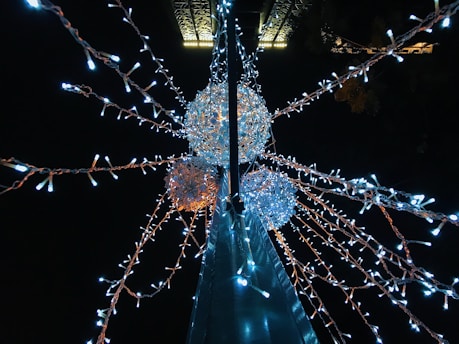 A night scene featuring a streetlight adorned with glowing white and orange string lights. The lights form a starburst pattern radiating from spherical structures, creating a festive and decorative atmosphere against the dark sky.