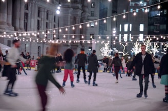 People are enjoying ice skating on a rink surrounded by festive string lights and illuminated trees. The scene is set in an urban area with large buildings in the background, and the atmosphere is lively with individuals of varying attire skating in different directions.