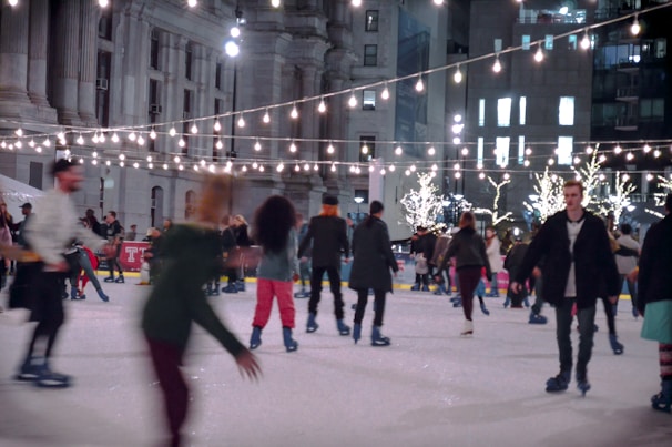 People are enjoying ice skating on a rink surrounded by festive string lights and illuminated trees. The scene is set in an urban area with large buildings in the background, and the atmosphere is lively with individuals of varying attire skating in different directions.