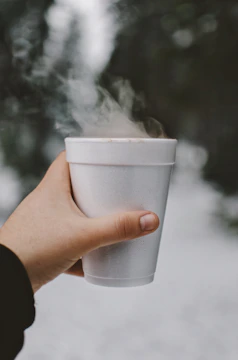 A hand holds a steaming white styrofoam cup against a blurred natural background. The steam rises visibly from the cup, suggesting a hot beverage inside.