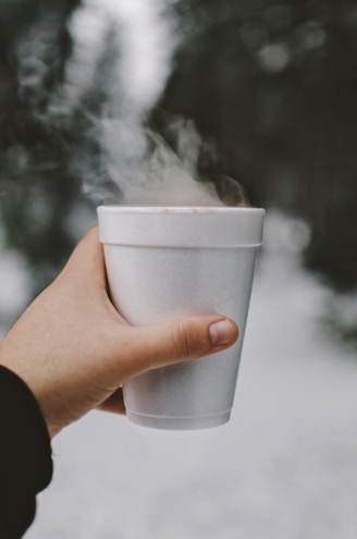Close-up of a hand holding a steaming cup of coffee next to a stack of local team hats.