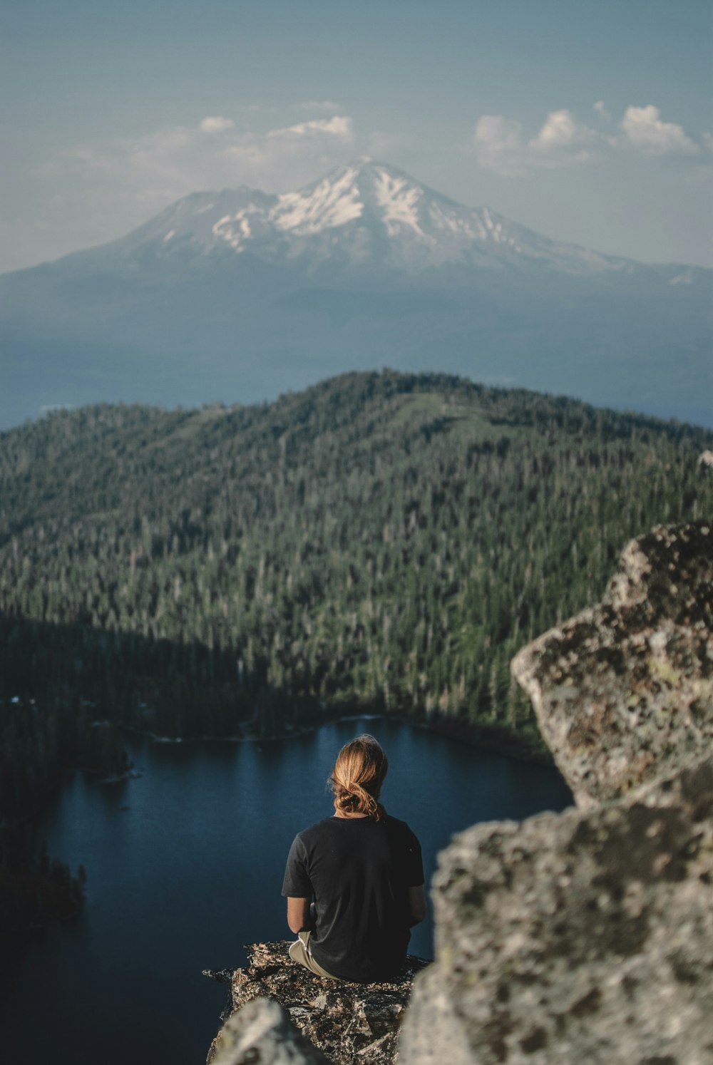 woman sitting on elevated ground overlooking body of water facing pine trees during daytime