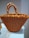 A large, intricately woven handbag featuring a crochet pattern in vibrant orange with yellow accents. The bag is placed on a white cloth, and the handles are wrapped with a textured, braided design.