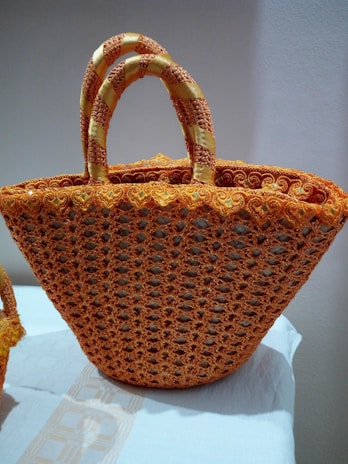A large, intricately woven handbag featuring a crochet pattern in vibrant orange with yellow accents. The bag is placed on a white cloth, and the handles are wrapped with a textured, braided design.