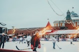 A group of people are playing ice hockey on an outdoor rink. The scene is decorated with string lights and festive elements, suggesting a holiday atmosphere. A historical building with red walls and towers is visible in the background, likely indicating a well-known location. The rink is surrounded by colorful advertisements and a few bystanders are watching the game.