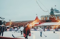 A group of people are playing ice hockey on an outdoor rink. The scene is decorated with string lights and festive elements, suggesting a holiday atmosphere. A historical building with red walls and towers is visible in the background, likely indicating a well-known location. The rink is surrounded by colorful advertisements and a few bystanders are watching the game.