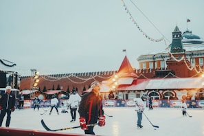 A group of people are playing ice hockey on an outdoor rink. The scene is decorated with string lights and festive elements, suggesting a holiday atmosphere. A historical building with red walls and towers is visible in the background, likely indicating a well-known location. The rink is surrounded by colorful advertisements and a few bystanders are watching the game.