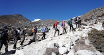 A group of people wearing hiking gear is trekking uphill on a rocky terrain with sparse vegetation. The sky is clear and blue, and mountains can be seen in the background. Each person carries a backpack and uses walking poles.