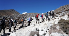 A group of people wearing hiking gear is trekking uphill on a rocky terrain with sparse vegetation. The sky is clear and blue, and mountains can be seen in the background. Each person carries a backpack and uses walking poles.