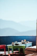 A tranquil outdoor setting features two small tables set up with ceramics and colorful dishes against a backdrop of blue mountains. Bright sunlight casts warm shadows across the vibrant rugs and furniture.